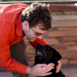 Photo of Dave wearing a red shirt and sunglasses bending over petting Katie the black lab guide dog in front of a brick wall