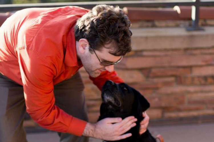 Photo of Dave wearing a red shirt and sunglasses bending over petting Katie the black lab guide dog in front of a brick wall