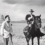 Black and White Photo: John Wayne Riding a horse wearing a cowboy hat and John Ford Standing next to him wearing a beret, white coat and using a cane in the desert with a few trees in the background