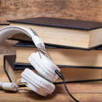 Stack of 3 books with black covers with a white pair of headphones next to them on a wood table