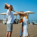 Set on a large wooden dock, overlooking the clear sky and ocean. Woman with long, blonde hair wearing sunglasses and a long, white vest over a yellow t-shirt and black shorts. Taller man with long, curly, brown hair wearing sunglasses and a white t-shirt with gray shorts. Woman is gesturing a "stop" symbol close to the man's face.