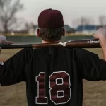 Young baseball player stands with his back to the camera, a baseball bat straddled on his shoulders with his arms slung over each end casually.